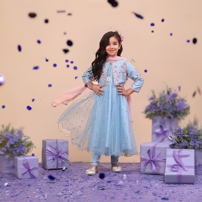 A girl wearing a blue net Anarkali suit set with printed kurta and straight sleeves, paired with matching churidar bottoms and a dupatta, standing in front of gift boxes and floral decor.